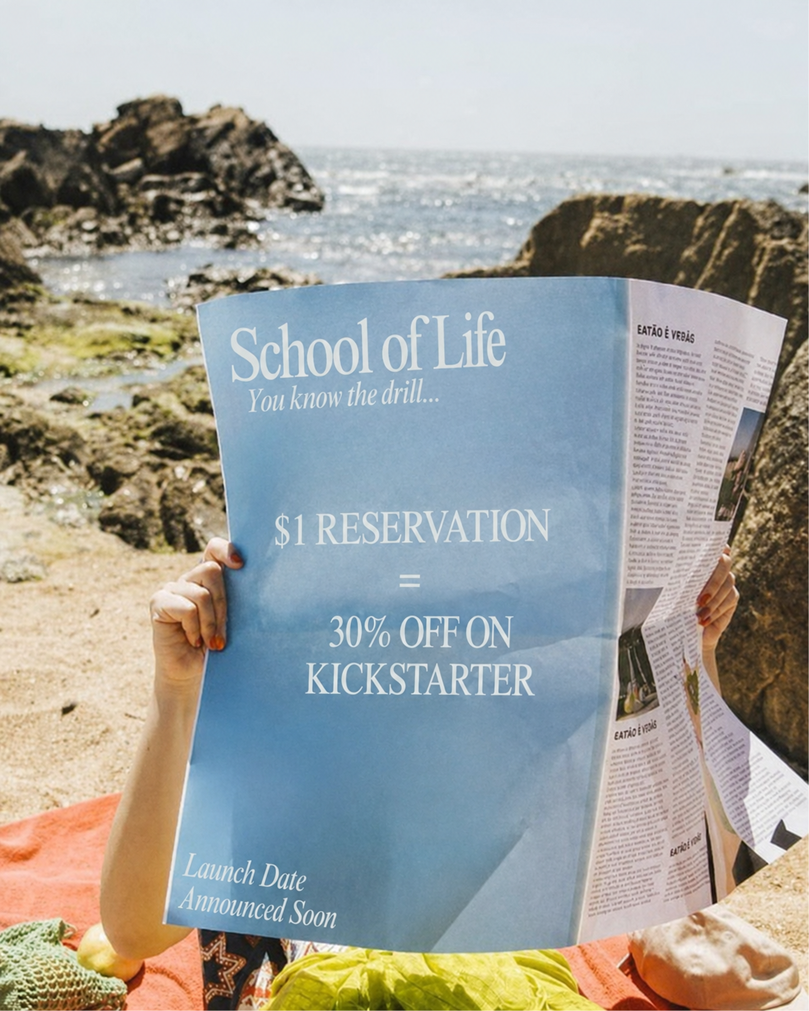 A person on a rocky beach holds a large blue printed poster or book cover in front of their face. The printed item reads 'School of Life You know the drill.